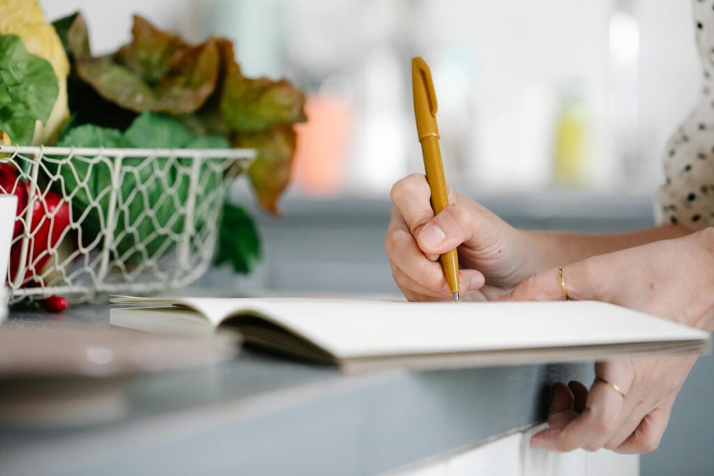 Close-up of a woman writing in a notebook on a kitchen counter beside a basket of fresh vegetables.