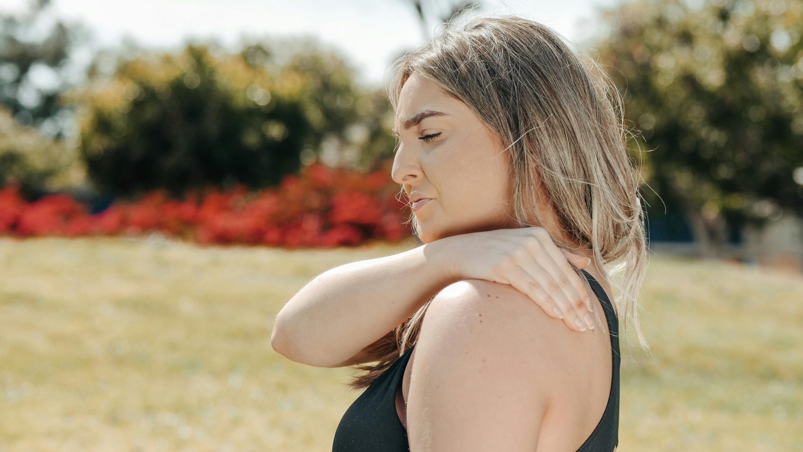 Woman in black tank top touching shoulder outdoors. Concept of shoulder pain and relaxation.